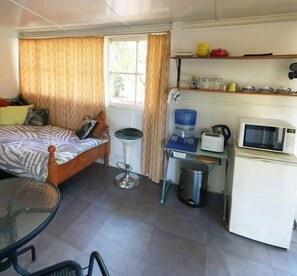 Habitación compacta con cama, una mesita, una silla, microondas, dispensador de agua y basurero.