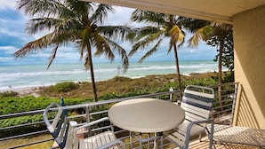 A balcony with a table and chairs overlooking the ocean, palm trees, and a beach. - Oceanfront Oasis – Longboat Key 226 by RVA (Longboat Key)