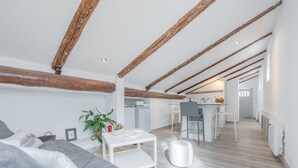 Un loft moderne avec des poutres apparentes, un canapé gris, une petite table blanche et un coin cuisine à l’arrière-plan.
