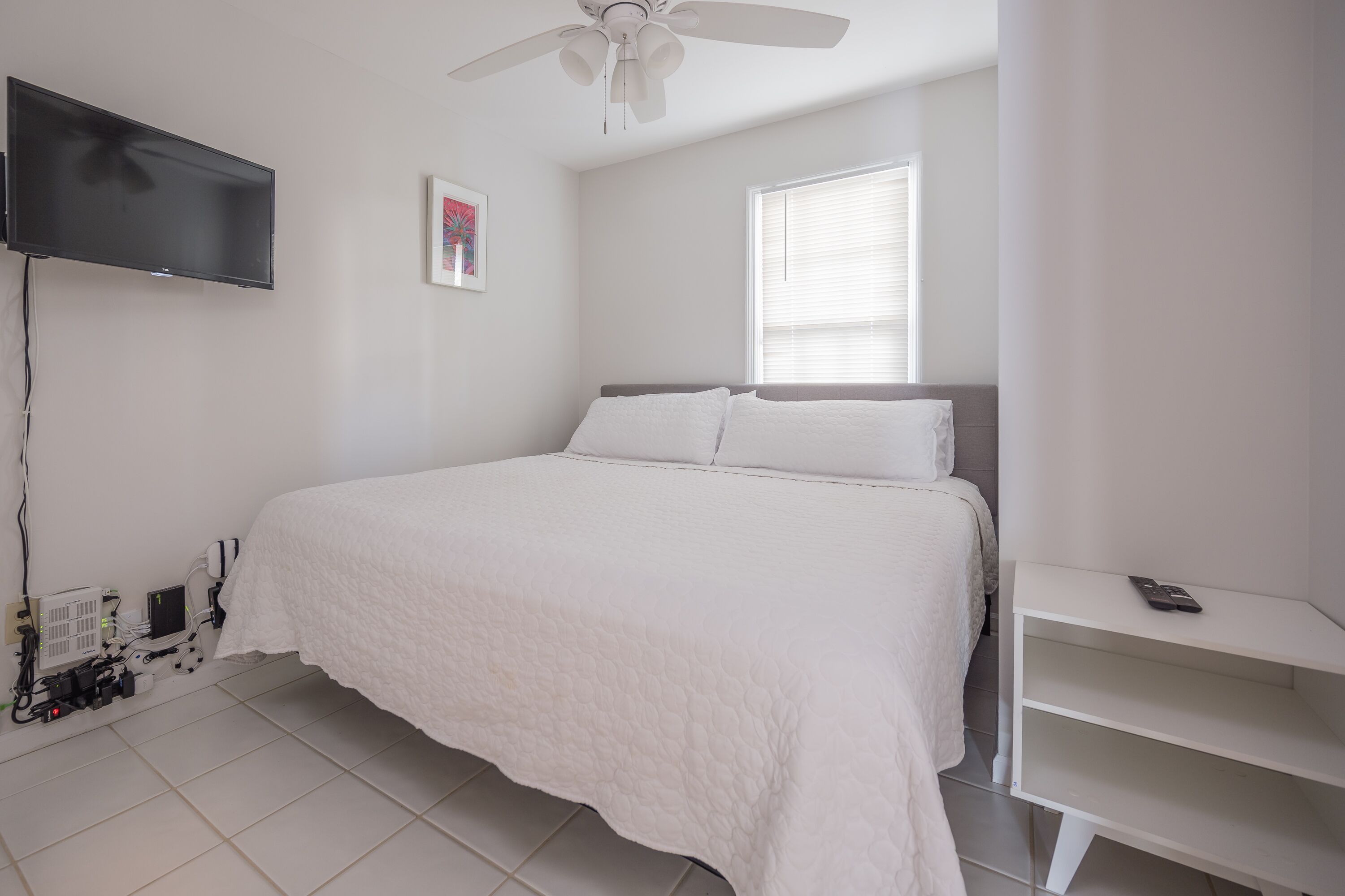 Une chambre à coucher avec un lit, un téléviseur fixé au mur, un ventilateur de plafond et une fenêtre avec des stores.