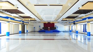 Ein geräumiger, moderner Saal mit glänzendem Boden, einer Farbgestaltung in Blau und Gelb sowie einer zentralen Bühne mit rotem Vorhang.