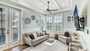 Ein großzügiges Wohnzimmer mit grauem Sofa, einem kleinen Sessel, einem Couchtisch und einem Deckenventilator. Große Fenster mit Jalousien sorgen für natürliches Licht, und ein Flachbildfernseher ist an der Wand befestigt.