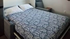 A small, compact room with a bed, a chair, a window, and a wall-mounted shelf. - Studio Condo (Newly Renovated) - 150' to Beach 20 min to Portland (Scarborough)