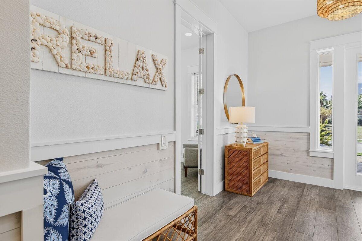 A room with a wooden floor, a white wall with a 'RELAX' sign, a wicker chair, a wooden side table with a lamp, and a view of the outdoors through a window.