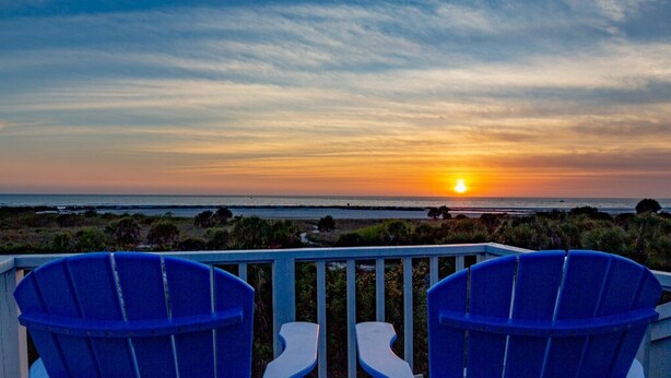 Une vue sur le coucher de soleil depuis un balcon, avec deux chaises bleues donnant sur l’océan.