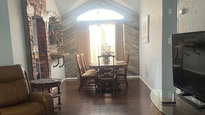 A living room with a brown leather sofa, a glass coffee table, and a TV on a wooden stand. There is a dining area with a table and chairs, and a brick wall with decorative items. - Spacious 4BR Retreat with Pool and Spa (Rowlett)