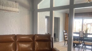 A spacious living room with a brown leather sofa, a glass coffee table with a potted plant, and a dining area in the background. - Spacious 4BR Retreat with Pool and Spa (Rowlett)