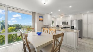 Una cocina moderna con armarios blancos, una isla grande y una zona de comedor con vista a palmeras.