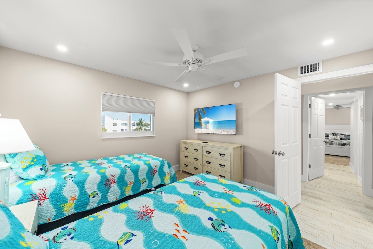 A bedroom with two beds, a ceiling fan, a TV, and a view of the beach through a window.