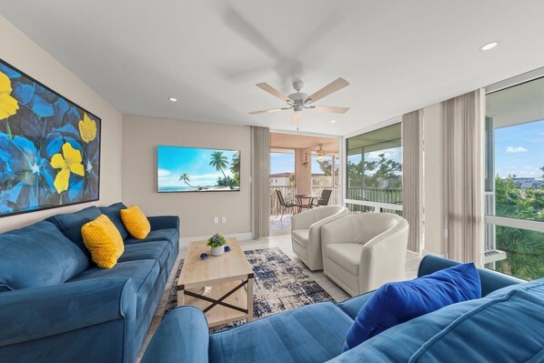 A modern living room with a blue sofa, a coffee table, and a ceiling fan. There is a large window with a view of palm trees and a balcony.
