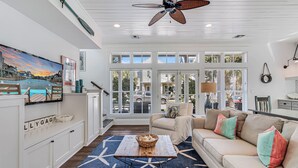 A spacious living room with a large TV, a ceiling fan, and a view of the outdoors through large windows.