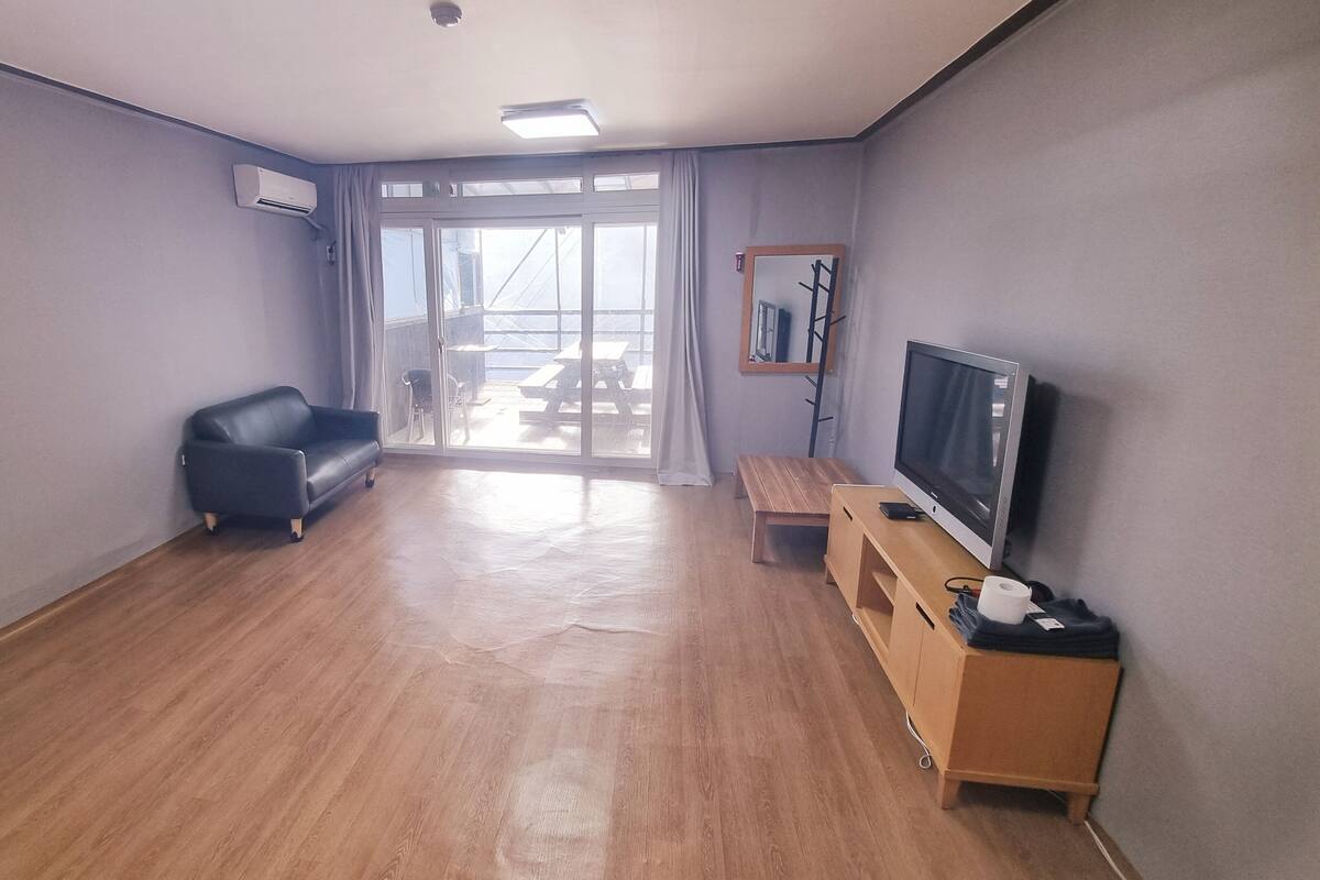 Ein Zimmer mit Holzboden, einem schwarzen Ledersofa, einer Holzbank, einem Fernseher auf einem Holzständer und einer Glastür, die zu einem Außenbereich führt.