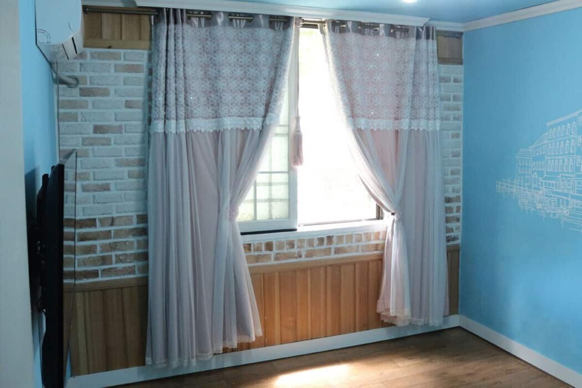 Ein Zimmer mit Backsteinwand, einem Fenster mit durchsichtigen Vorhängen, Holzboden und einer blauen Wand mit Gebäudeumriss.