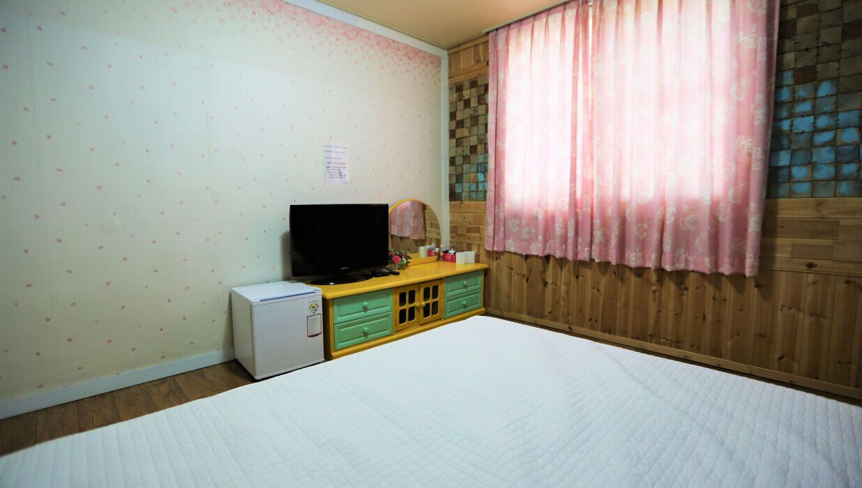 A bedroom with a bed, a small desk with a computer, a television, and a window with curtains.