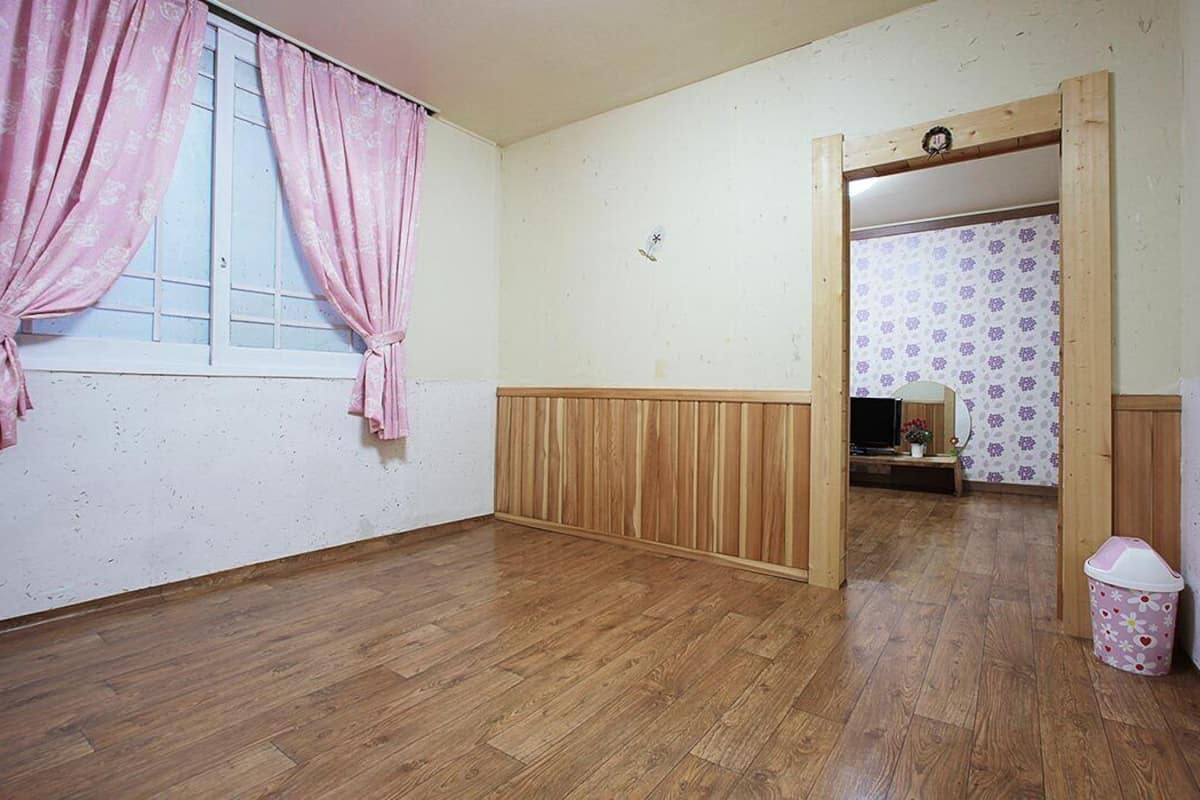Ein Zimmer mit Holzboden, einer Wandpaneele aus Holz, einem Fenster mit rosa Vorhängen und einer Tür zu einem weiteren Raum mit gemustertem Wandpapier.