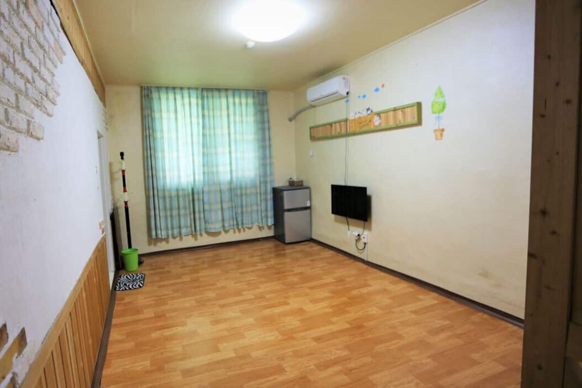 Ein Zimmer mit Holzboden, Ziegelwand, Fenster mit Vorhängen, einem kleinen Kühlschrank und einem Wand-Fernseher.