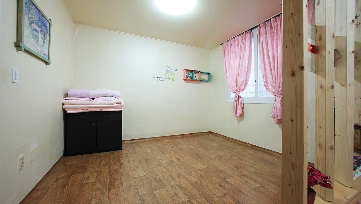 A room with wooden flooring, a black wardrobe, a small shelf with items, and pink curtains on a window.