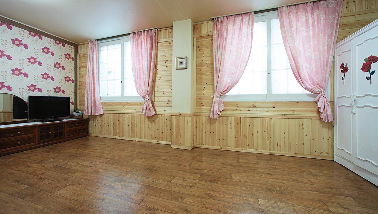 Una habitación con paneles de madera, un televisor sobre un mueble de madera y cortinas rosadas en las ventanas.