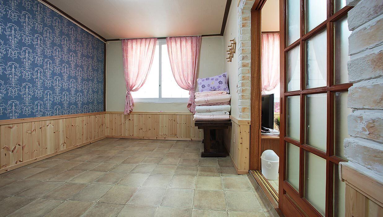 A room with a wooden floor, a window with pink curtains, a bed with a floral pillow, and a brick wall.