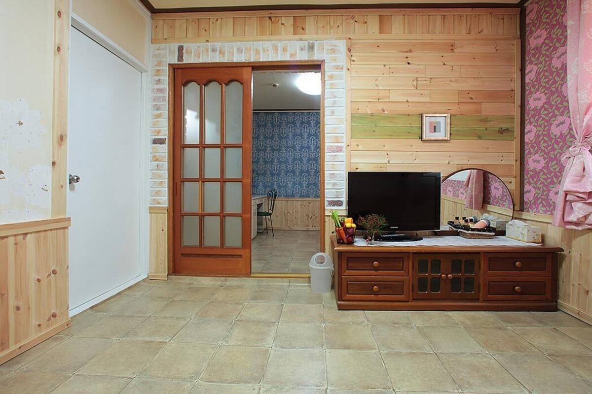 Ein Zimmer mit Holzboden, einem Fernseher auf einem Holzkasten, einer Tür zu einem anderen Raum und einem Fenster mit Vorhängen.