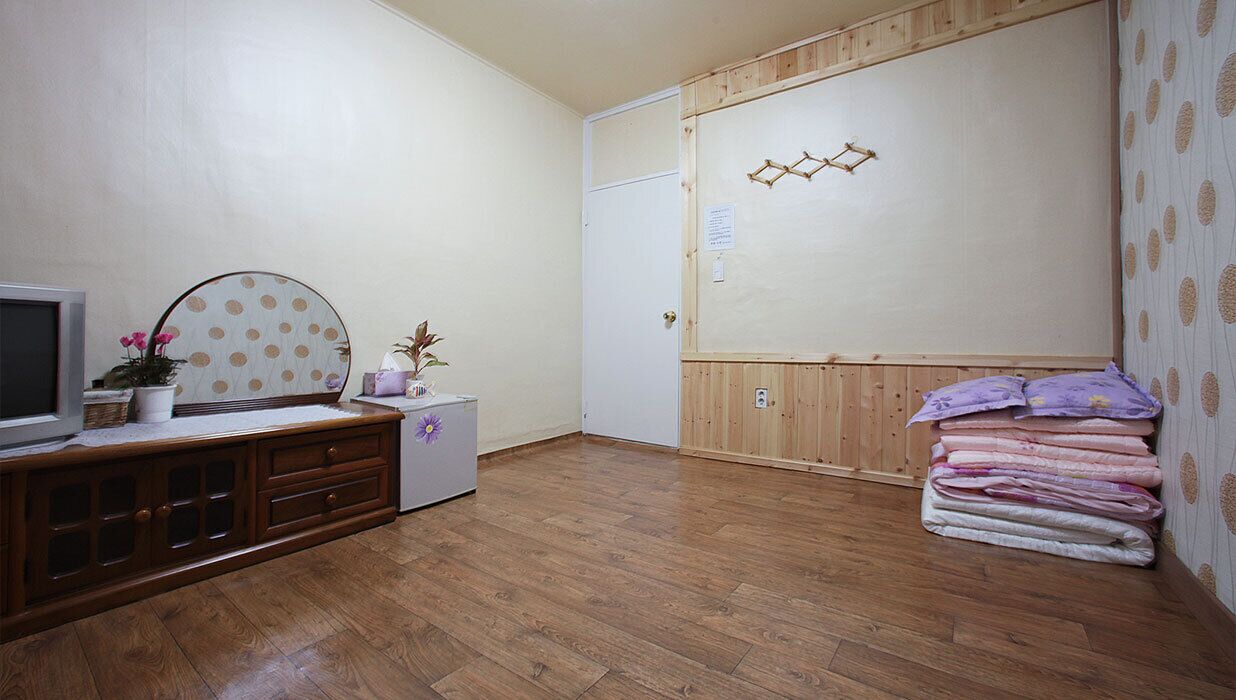 Una habitación con piso de madera, un televisor sobre un mueble, un refrigerador pequeño y una cama con mantas dobladas.