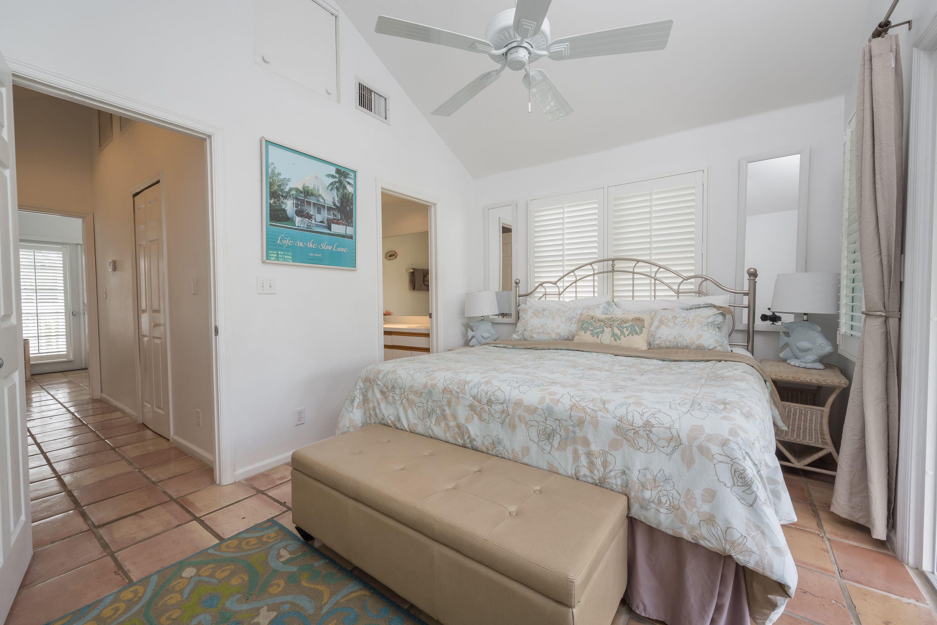 A bedroom with a bed, bench, ceiling fan, and a door leading to another room.