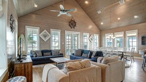 A spacious living room with a high ceiling, wooden beams, and large windows. There is a dining area with a table and chairs, and a kitchen area with a countertop and cabinets.