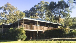 A two-story wooden house with a balcony, surrounded by trees and a grassy area.