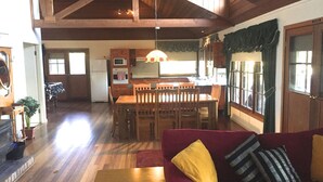 A spacious living room with a red sofa, wooden dining table, and chairs. A kitchen area with wooden cabinets and a hanging light fixture is visible in the background.