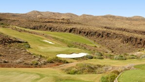 A golf course with a bunker, green areas, and rocky hills in the background.