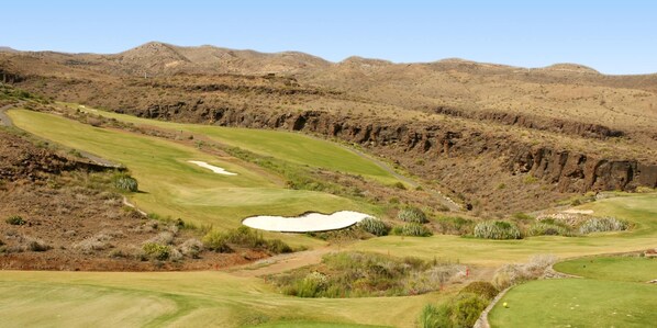 Un campo de golf con un búnker, zonas verdes y colinas rocosas al fondo.