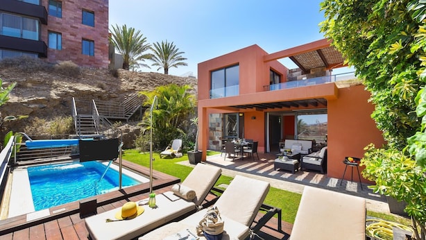 A modern outdoor pool area with lounge chairs, a barbecue, and a two-story building with large windows.