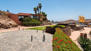 A modern building with a clear view of the surrounding landscape, including a paved area with bollards, a landscaped garden with red flowers, and a distant view of the ocean.