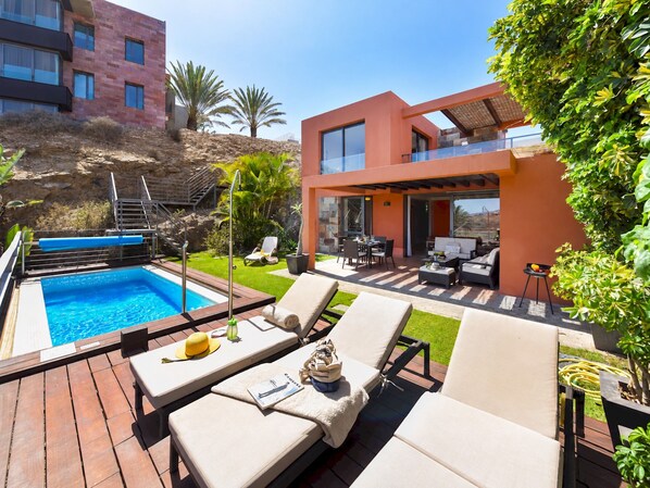 A modern outdoor pool area with lounge chairs and a hot tub, surrounded by a landscaped garden and a two-story building.