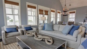 A spacious living room with a large gray sectional sofa, two armchairs, a coffee table with a unique sculpture, and a dining area in the background. - Stunning Designer Home w/ Pool Views+Access to Cinnamon Shore Amenities (Port Aransas)