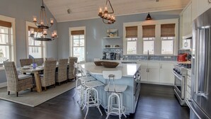 A modern kitchen with a central island, bar stools, and a dining area with wicker chairs. - Stunning Designer Home w/ Pool Views+Access to Cinnamon Shore Amenities (Port Aransas)
