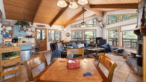A spacious living area with a high ceiling, wooden beams, and large windows offering views of the outdoors.