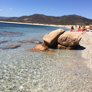 En strand med klart vatten, stora stenar och människor i bakgrunden.