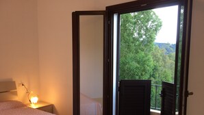 A bedroom with a bed, a nightstand with a lamp, and a balcony with a view of trees. - Charming apartment on the mountainside near the sea (Villaputzu)
