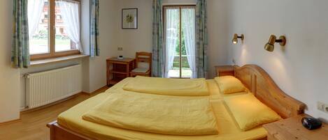 A bedroom with a large bed, a wooden nightstand, a chair, a desk, and a window with curtains.