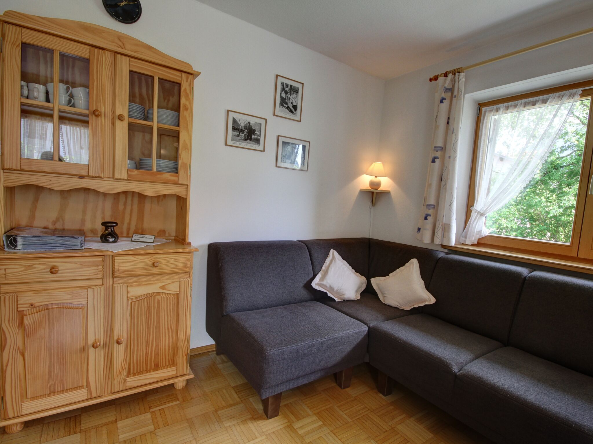 A cozy living room with a dark grey sofa, wooden cabinet with glass doors, and a window with curtains.