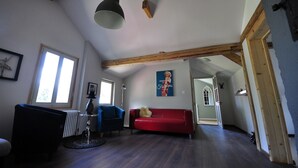 Ein geräumiges Wohnzimmer mit einem roten Sofa, einem blauen Sessel und einem schwarzen Stuhl. Ein Holzbalken verläuft an der Decke und eine schwarze Pendelleuchte ist angebracht. Ein gerahmtes Bild hängt an der Wand, ein Poster ist sichtbar.