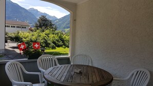Ein Balkon mit Holztisch und weißen Stühlen, mit Blick auf eine Berglandschaft und einer Schweizer Flagge.