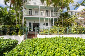 A two-story building with a balcony, surrounded by lush greenery and a white fence.