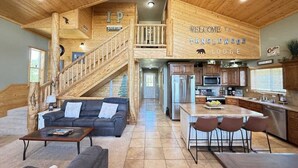 A spacious lodge interior with a wooden staircase leading to a loft, a cozy living area with a sofa and coffee table, a kitchen with modern appliances, and a dining area with a large island.