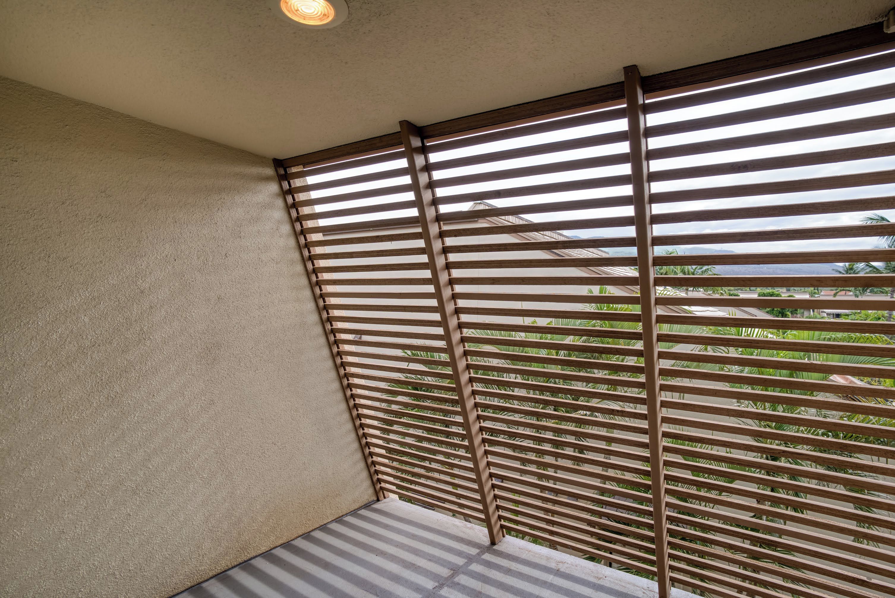 Un coin de pièce avec un mur beige à la texture particulière, une cloison en bois à lattes et, en arrière-plan, une vue sur de la verdure et un bâtiment.