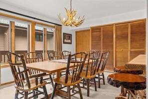 A dining area with a wooden table and chairs, a chandelier, and a view through large windows.