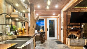 A rustic kitchen with a stone floor, wooden shelves, and a large TV on the wall. - Cedar Springs Cabin I Putting Green + Hot Tub (Hot Springs)