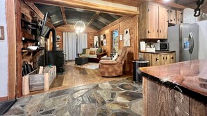 A rustic cabin interior with a stone floor, wooden walls, a fireplace, a sofa, a dining table, and a kitchen area with a refrigerator and microwave. - Cedar Springs Cabin I Putting Green + Hot Tub (Hot Springs)