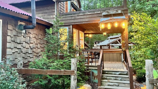 A rustic wooden house with a wooden staircase leading to a porch with hanging lights. - Cedar Springs Cabin I Putting Green + Hot Tub (Hot Springs)